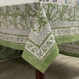 May include: A green and white floral patterned tablecloth. The tablecloth features a repeating pattern of birds, flowers, and leaves. The border has a decorative design. The tablecloth is draped over a wooden table.