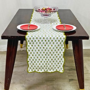 Peut inclure: Une table de salle à manger dressée. La table a une surface marron foncé avec un chemin de table blanc à motifs floraux verts. Deux assiettes rouges avec des fourchettes sont de chaque côté du chemin de table. Un bol en verre avec des objets décoratifs est au centre. Les pieds de la table ont des accents dorés.