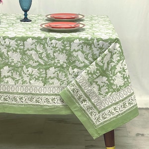 May include: A light green tablecloth featuring a white floral and bird pattern with a decorative border. The fabric is draped over a table set with two plates, each holding a silver fork, and a blue glass goblet positioned on the surface.
