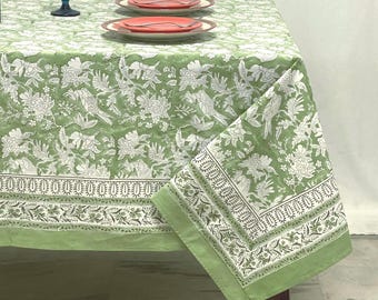 Puede incluir: Un mantel verde claro con un patrón floral y de aves en blanco con un borde decorativo. La tela cubre una mesa dispuesta con dos platos, cada uno con un tenedor de plata, y una copa de cristal azul colocada sobre la superficie.
