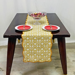 Peut inclure: Une table à manger en bois foncé est ornée d'un chemin de table à motifs jaunes et blancs, de deux assiettes rouges et d'un bol en verre rempli d'objets décoratifs. Les pieds de la table ont des accents dorés. Le texte "FLORALPRINT .CO" est visible.