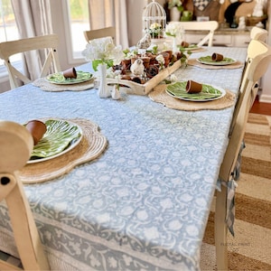May include: A dining table covered with a light blue and white floral patterned tablecloth. The table is set with round woven placemats, green leaf-shaped plates, and small brown terracotta pots. A wooden centerpiece holds more pots.
