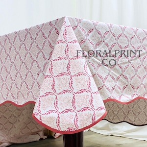 Puede incluir: Un mantel estampado en rosa y blanco con un borde festoneado. El mantel presenta un diseño floral repetido y el texto "FLORAL PRINT CO". El mantel está drapeado sobre una mesa.