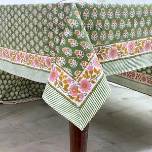 Puede incluir: Un mantel floral verde con un patrón repetido de pequeñas flores y un borde de flores rosas y amarillas más grandes. El mantel está sobre una mesa con patas de color marrón oscuro.