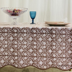 Mud Brown floral cotton tablecloth photographed on a long rectangular dining table with rustic farmhouse décor accents. The handcrafted hand block print design and scalloped piping detail highlight artisan craftsmanship while offering a stylish reusable table cover suitable for parties, family dinners, wedding events, holiday gatherings, and cozy modern boho interiors.