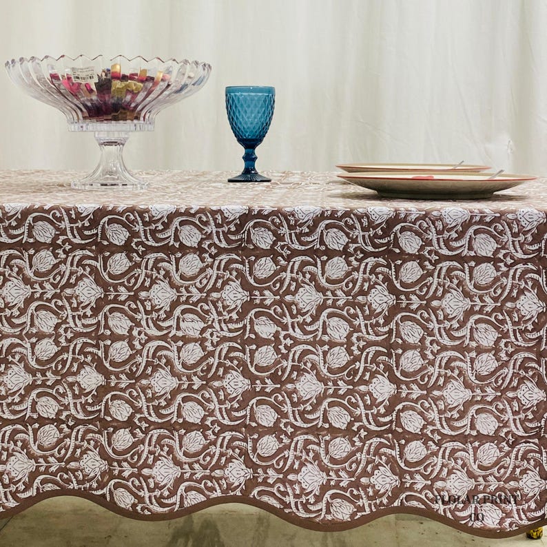 Mud Brown floral cotton tablecloth photographed on a long rectangular dining table with rustic farmhouse décor accents. The handcrafted hand block print design and scalloped piping detail highlight artisan craftsmanship while offering a stylish reusable table cover suitable for parties, family dinners, wedding events, holiday gatherings, and cozy modern boho interiors.