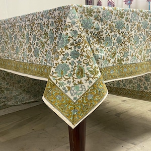 May include: A white tablecloth with a floral pattern in shades of blue, green, and brown. The border features a repeating design in gold and green. The tablecloth is draped over a wooden table, showcasing its intricate details and design.
