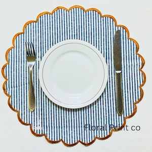 May include: A round placemat with a blue and white vertical striped pattern and a scalloped edge trimmed in orange. Centered on the placemat is a white plate with a gold rim, flanked by a silver fork on the left and a silver knife on the right.