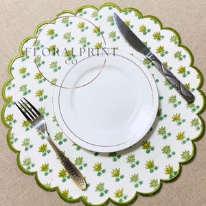 May include: A white placemat with a green scalloped edge and a floral print, featuring a white plate, a fork, and a knife. The text "FLORAL PRINT CO" is printed on the placemat.