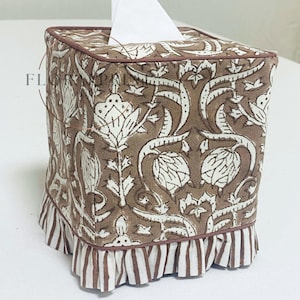 May include: A square tissue box cover with a brown and white floral pattern. The cover has a brown trim and a striped ruffle at the base. White tissues are visible at the top. The design is reminiscent of vintage textiles.