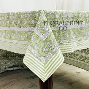 Puede incluir: Un mantel con estampado floral verde y blanco con el texto "FLORALPRINT CO". El mantel presenta un diseño floral repetido y un borde a rayas. La esquina del mantel está drapeada sobre una pata de mesa marrón oscuro.