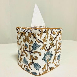 Puede incluir: Una funda de caja de pañuelos cuadrada con un estampado floral en tonos azules, marrones y dorados sobre fondo blanco. Tiene un ribete marrón y un pañuelo blanco visible en la parte superior.