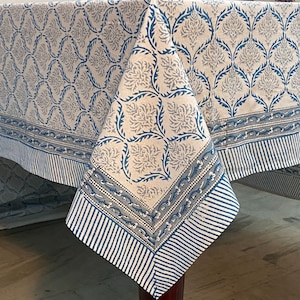 Puede incluir: Un mantel blanco con un estampado floral azul y gris. El mantel tiene un borde con rayas azules verticales y una banda decorativa. La tela está doblada sobre una mesa.