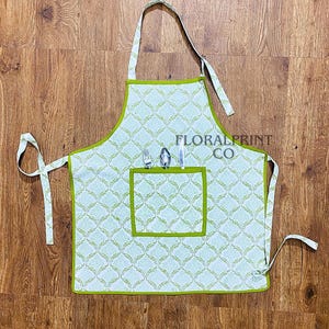 Może przedstawiać: Jasnoniebieski i biały wzorzysty fartuch z zielonym wykończeniem i kieszenią. Fartuch ma kwiatowy wzór i napis "FLORAL PRINT CO". W kieszeni znajdują się widelec i nóż.