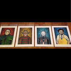 May include: Four framed prints of horror movie characters. Each print features a different character in a colorful, stylized illustration against a patterned background. The characters are Jason Voorhees, Freddy Krueger, Pinhead, and Leatherface.