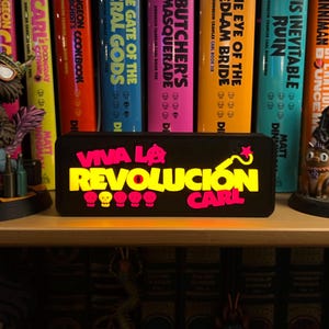 May include: A shelf displays colorful books and figurines. The books have titles like "Dungeon Crawler" and "The Butcher's Masquerade." A lighted sign reads "Viva La Revolución Carl." Figurines include a dinosaur and a character with a dog.