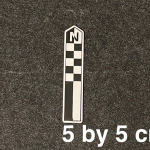 K&ouml;nnte beinhalten: Ein schwarz-wei&szlig;er Kompassrosen-Aufkleber, 5 cm x 5 cm gro&szlig;. Der Aufkleber hat ein Schachbrettmuster und den Buchstaben "N" oben, der Norden anzeigt. Der Aufkleber befindet sich auf einem dunkelgrauen Hintergrund.