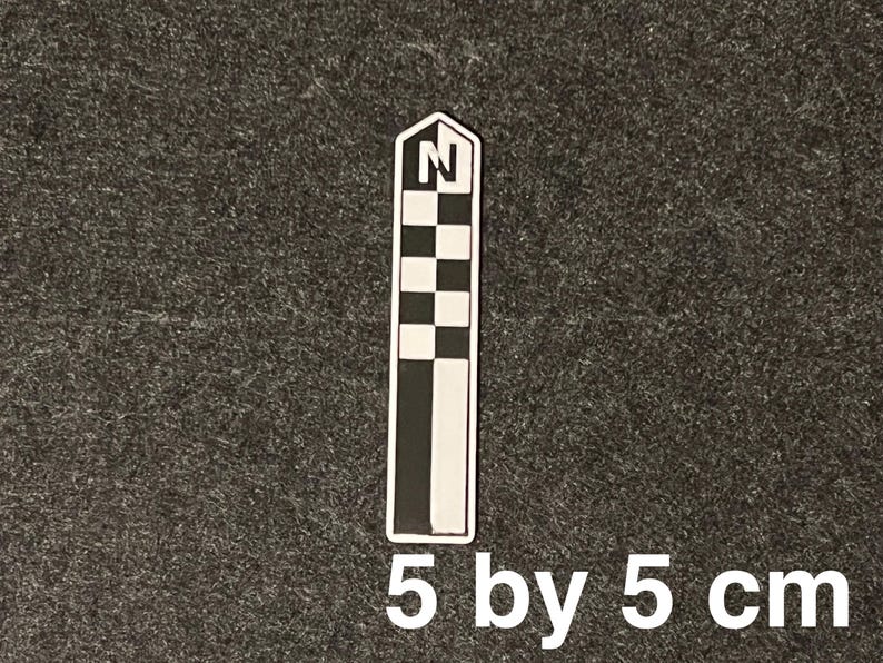 K&ouml;nnte beinhalten: Ein schwarz-wei&szlig;er Kompassrosen-Aufkleber, 5 cm x 5 cm gro&szlig;. Der Aufkleber hat ein Schachbrettmuster und den Buchstaben "N" oben, der Norden anzeigt. Der Aufkleber befindet sich auf einem dunkelgrauen Hintergrund.