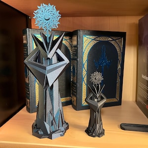 May include: Two geometric sculptures in shades of gray and blue, resembling stylized figures with floral accents. The larger sculpture is approximately 8 inches tall, while the smaller one is about 4 inches. They are displayed on a wooden shelf with decorative books.