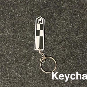 K&ouml;nnte beinhalten: Ein rechteckiger Schl&uuml;sselanh&auml;nger mit einem schwarz-wei&szlig;en Schachbrettmuster und dem Buchstaben "N" in einem wei&szlig;en Feld. Er ist an einem silbernen Metallring befestigt. Das Wort "Keychain" ist in wei&szlig;er Schrift geschrieben.
