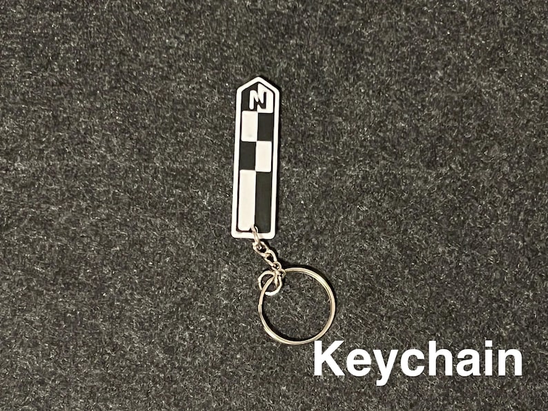 K&ouml;nnte beinhalten: Ein rechteckiger Schl&uuml;sselanh&auml;nger mit einem schwarz-wei&szlig;en Schachbrettmuster und dem Buchstaben "N" in einem wei&szlig;en Feld. Er ist an einem silbernen Metallring befestigt. Das Wort "Keychain" ist in wei&szlig;er Schrift geschrieben.