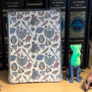 May include: A clear protective tablet case featuring a repeating pattern of fantasy-themed illustrations including hooded figures, swords, spears, books, potion bottles, and coins in shades of blue, tan, and red against a cream-colored background.