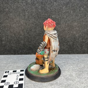 Op de afbeelding: Handgeschilderd beeldje van een personage met rood haar, met een cape met vleugelachtige details. Het figuur houdt een tas vast en staat op een cirkelvormige basis met gras en een steen. Het beeldje is ongeveer 5 cm hoog.