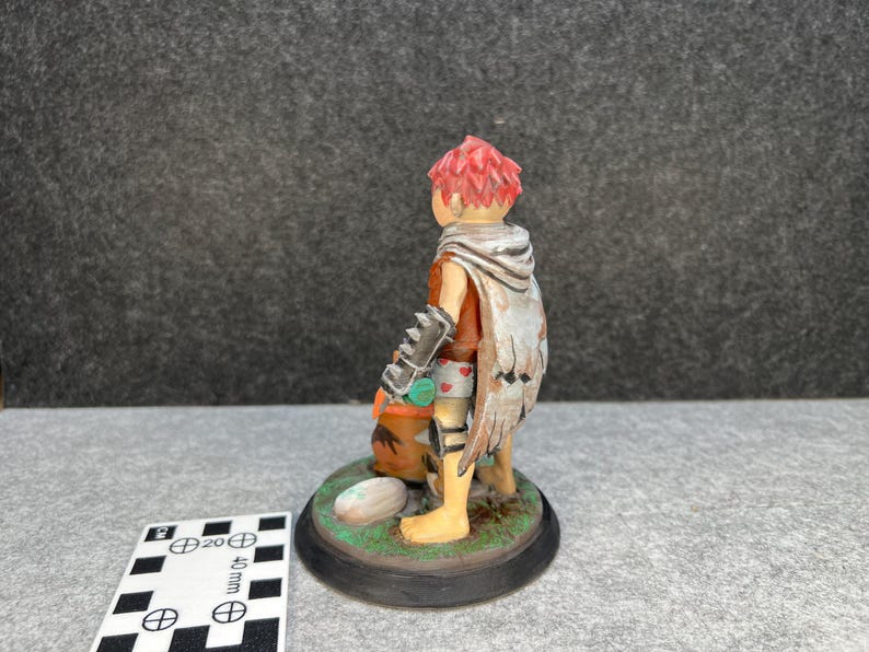 Op de afbeelding: Handgeschilderd beeldje van een personage met rood haar, met een cape met vleugelachtige details. Het figuur houdt een tas vast en staat op een cirkelvormige basis met gras en een steen. Het beeldje is ongeveer 5 cm hoog.