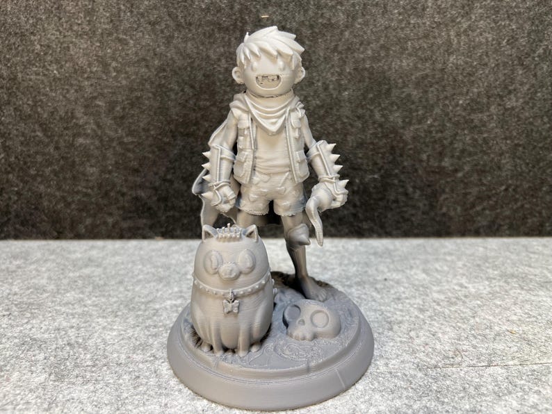 Op de afbeelding: Een grijze 3D-geprinte figuur van een jong personage met stekelige handschoenen, die een gebogen mes vasthoudt. De figuur staat op een cirkelvormige basis met een schedel en een katachtig wezen. Fantasiethema.