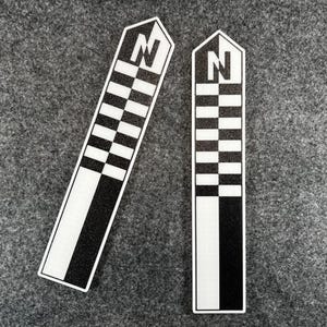K&ouml;nnte beinhalten: Zwei rechteckige Aufkleber mit einem schwarz-wei&szlig;en Schachbrettmuster und einem wei&szlig;en Rand. Die Oberseite jedes Aufklebers hat eine wei&szlig;e Form mit dem Buchstaben "N" in Schwarz. Die Aufkleber befinden sich auf einer grauen Oberfl&auml;che.