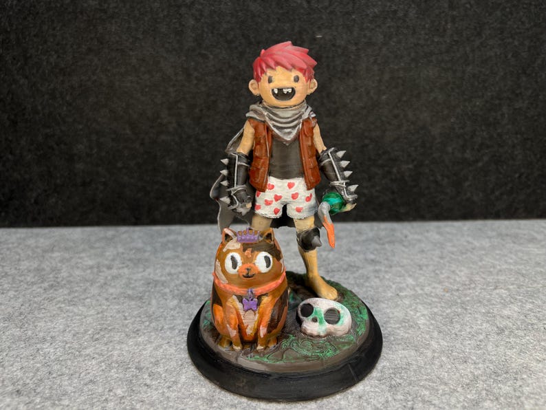 Op de afbeelding: Gedetailleerd beeldje van een stripfiguur met rood haar, een bruin vest, een korte broek met hartjespatroon en handschoenen met spikes. Een kat met een kroon en een schedel maken ook deel uit van het beeldje.