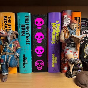 May include: A bookshelf display featuring books titled The Butcher's Masquerade, This Inevitable Ruin, and A Parade of Horribles by Matt Dinniman. Between the books stands a black rectangular object with four bright pink pixelated skull icons.