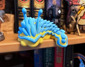 Könnte beinhalten: Eine 3D-gedruckte Meeresschnecken-Figur mit einer leuchtend gelben Basis und kräftig blauen Rückenanhängen. Das Wesen hat zwei markante blaue Fühler, die nach oben ragen, sowie einen texturierten, segmentierten Körper mit einer glatten gelben Unterseite.