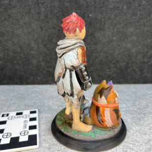 Op de afbeelding: Handgeschilderd beeldje van een persoon met rood haar en een witte en grijze mantel, naast een katachtig wezen. Het beeldje staat op een zwarte basis met een groene rand. Een wit en zwarte meetkaart staat op de voorgrond.