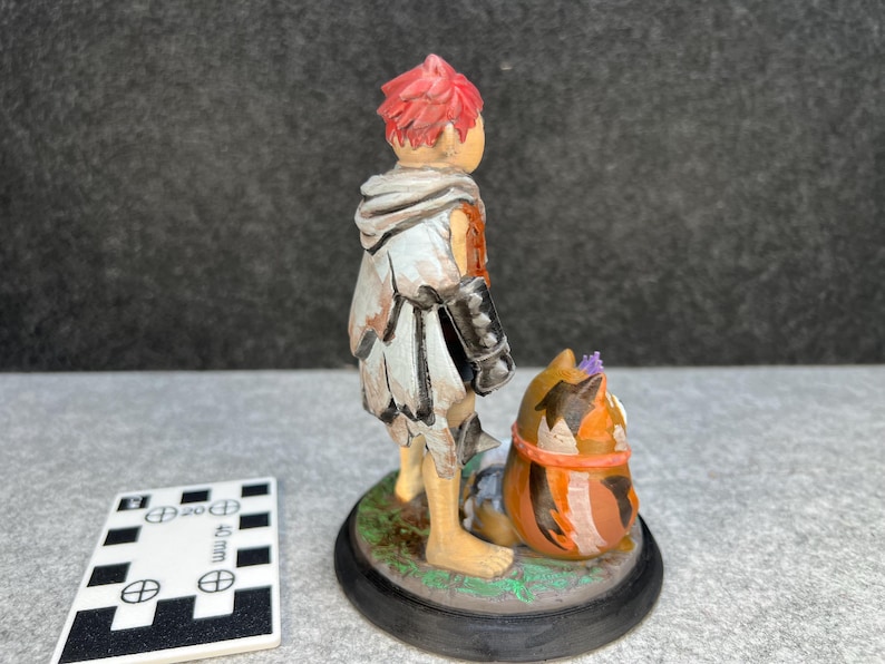 Op de afbeelding: Handgeschilderd beeldje van een persoon met rood haar en een witte en grijze mantel, naast een katachtig wezen. Het beeldje staat op een zwarte basis met een groene rand. Een wit en zwarte meetkaart staat op de voorgrond.