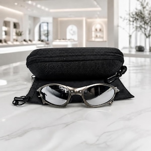May include: A pair of silver-toned metal sunglasses with reflective lenses resting on a black fabric pouch in front of a black zippered hard-shell protective case. The sunglasses feature a distinct wrap-around frame design with visible metal rivets.
