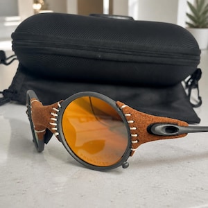 May include: Sunglasses with round orange polarized lenses and a matte black frame. The frame features brown suede-like material accents secured with visible white stitching around the lens edges. A black protective hard case is positioned behind.