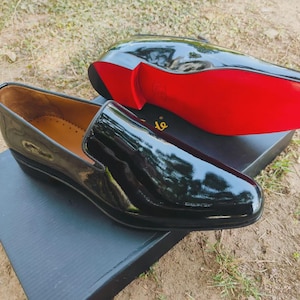 Puede incluir: Un par de mocasines negros de charol con acabado brillante. Un zapato está colocado sobre una caja negra, con el otro encima, mostrando una suela roja brillante. Los zapatos tienen un diseño clásico y elegante.