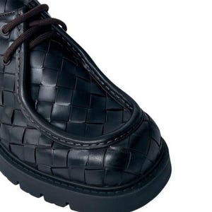 May include: Close-up of a black leather shoe with a woven pattern. The shoe features dark brown laces and a thick, black sole. The design showcases a unique texture and a modern aesthetic.