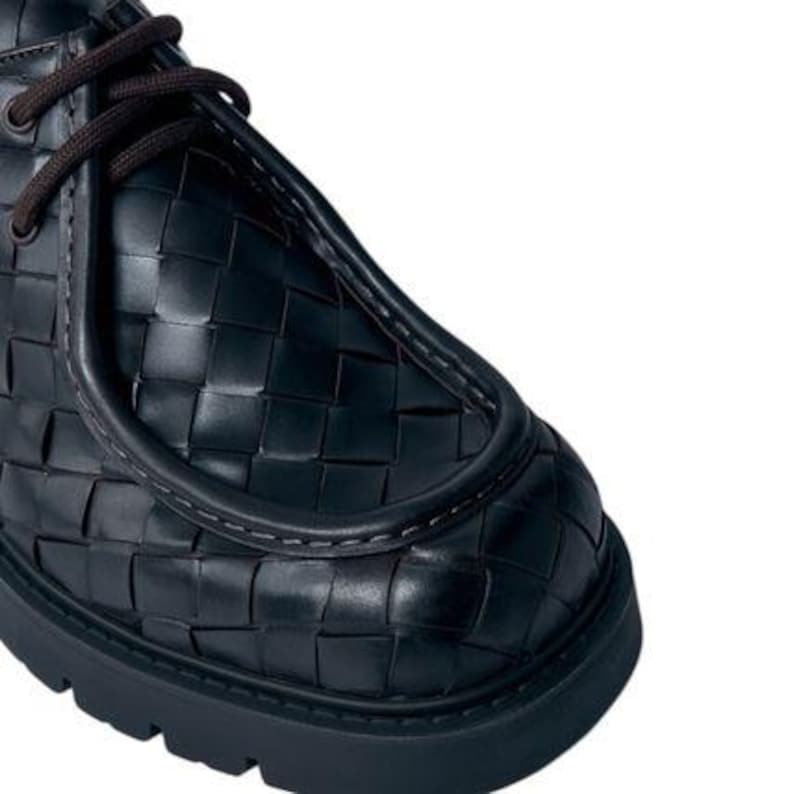 May include: Close-up of a black leather shoe with a woven pattern. The shoe features dark brown laces and a thick, black sole. The design showcases a unique texture and a modern aesthetic.