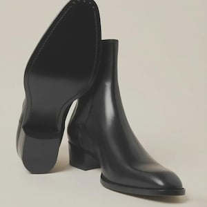 May include: A pair of black leather Chelsea boots with a smooth finish, featuring a rounded toe, a stacked block heel, and a clean, minimalist silhouette. One boot is positioned upright, while the other is tilted to reveal the smooth black sole.