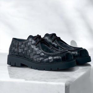 May include: A pair of black leather shoes with a woven pattern. The shoes have a thick sole and dark laces. The shoes are displayed on a white surface.