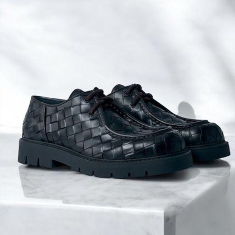 May include: A pair of black leather shoes with a woven pattern. The shoes have a thick sole and dark laces. The shoes are displayed on a white surface.