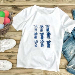 Może przedstawiać: Biały t-shirt z krótkim rękawem z centralnym nadrukiem graficznym przedstawiającym dziewięć niebieskich ananasów ułożonych w siatkę trzy na trzy. Ilustracje ananasów mają teksturowany, akwarelowy wygląd z różnymi odcieniami niebieskiego.