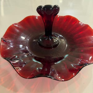 Puede incluir: Un plato de servicio de vidrio rojo intenso con un asa vertical central de remate redondeado. El plato tiene una base circular con un borde ondulado y festoneado, y un acabado translúcido que permite el paso de la luz a través del material.