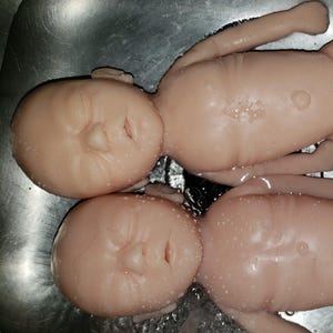 Może przedstawiać: Dwie bladoróżowe lalki dla dzieci są częściowo zanurzone w wodzie. Lalki leżą obok siebie, z widocznymi twarzami i ciałami. Lalki mają zamknięte oczy i szczegółowe rysy twarzy. Woda tworzy odblaskową powierzchnię.