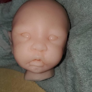 Pode incluir: Uma cabeça de boneca rosa pálido com olhos fechados, um nariz pequeno e uma boca ligeiramente aberta. A cabeça é feita de um material liso e tem uma base para o pescoço. A cabeça está sobre uma toalha verde.