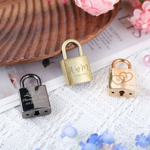 Könnte beinhalten: Drei kleine Vorhängeschlösser in Gold und Schwarz. Die goldenen Vorhängeschlösser tragen die eingravierten Initialen "A ♡ M" und "N ♡ K" sowie Daten. Das schwarze Vorhängeschloss trägt die Namen "Harry & Rosa" und ein Datum. Alle haben gebogene Bügel.
