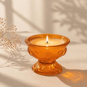 Pode incluir: Uma vela acesa em um castiçal de vidro âmbar. A vela está em um recipiente de vidro decorativo com base de pedestal. O vidro tem detalhes ornamentados. O fundo é de cor neutra com sombras.