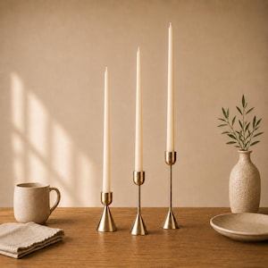 Può includere: Tre candele affusolate color crema in portacandele in ottone di diverse altezze. Una tazza beige, un tovagliolo di lino piegato, un piattino e un vaso strutturato con verde sono su un tavolo di legno.